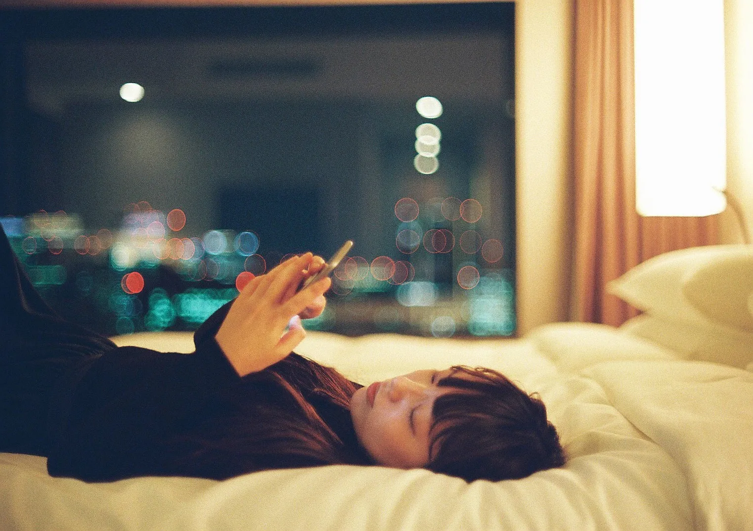 夜、ベッドに寝転んでスマートフォンを見る女性